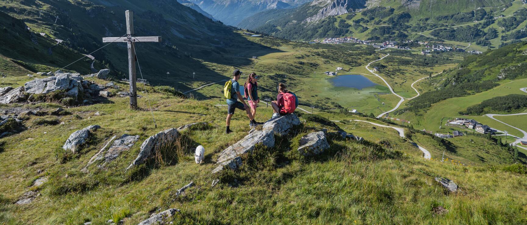 Three hikers with backpacks and dog at summit cross, view into green valley with lake and mountain village.