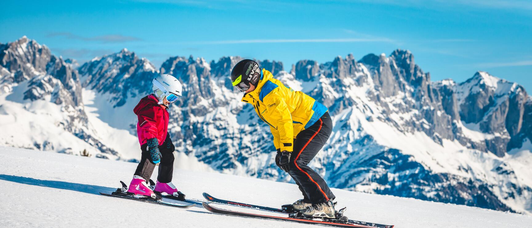 Ski instructor in yellow jacket teaching a young child on a snowy slope with rock peaks in the background.