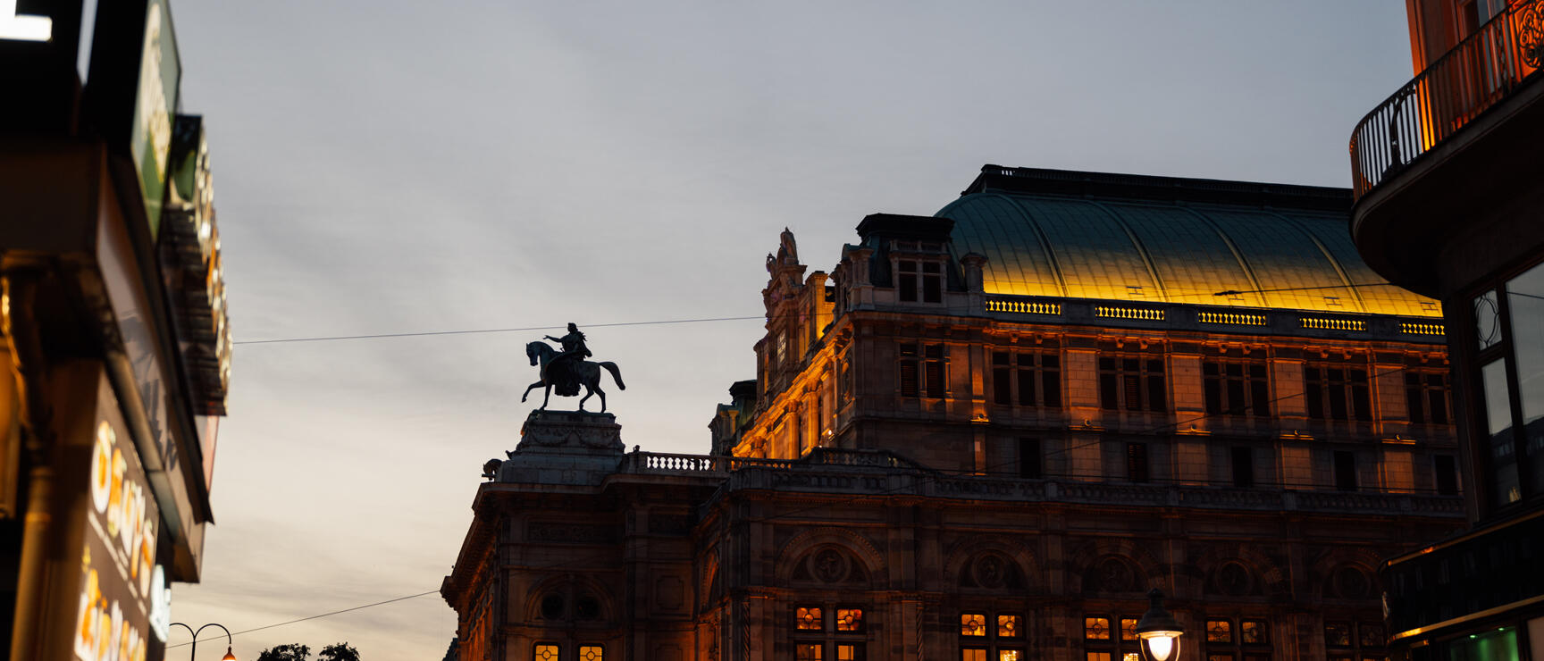 Vienna State Opera at dusk, illuminated, with equestrian statue in foreground.