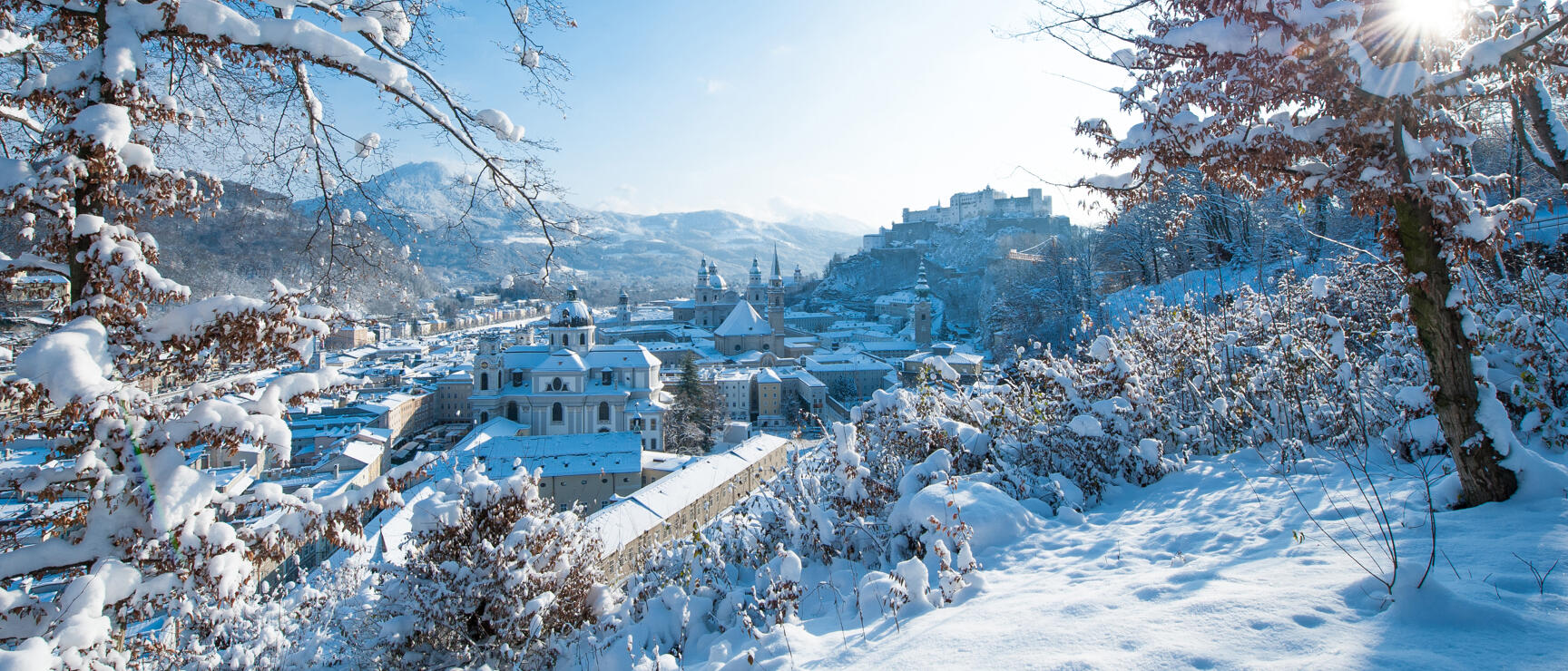 Snowy old town of Salzburg with Hohensalzburg Fortress, snow-covered trees in foreground, blue winter sky.