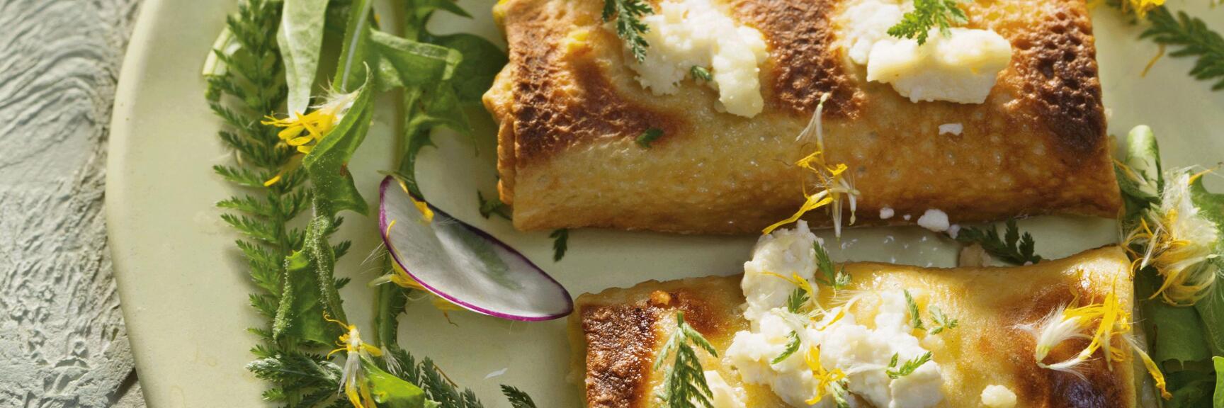 Two filled, fried palatschinken with cream cheese on plate with wild herbs, radish slices and yellow flowers.