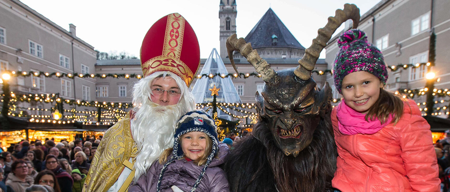 Father Christmas and Krampus with two children at Salzburg Christmas Market, illuminated market stalls in background.