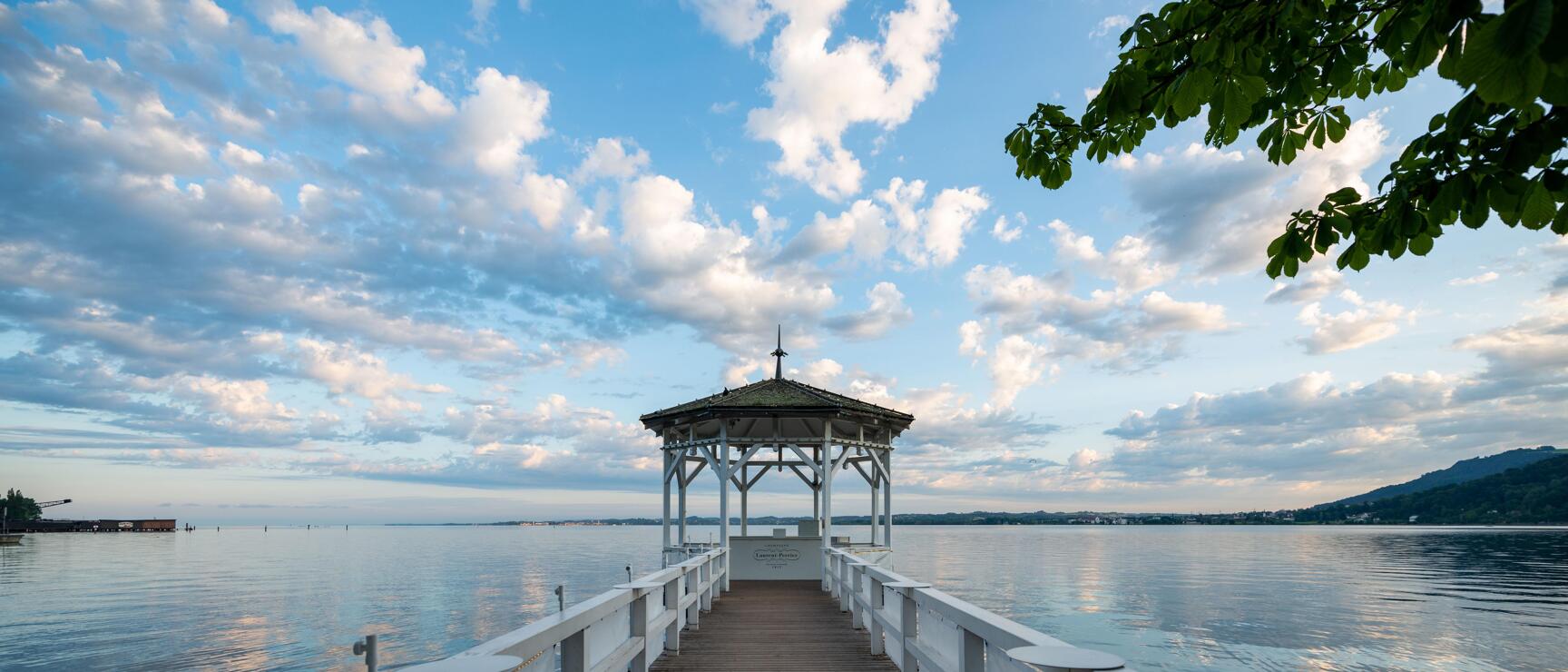White wooden pier with pavilion on Lake Wörthersee, calm water and cloudy sky in the background.