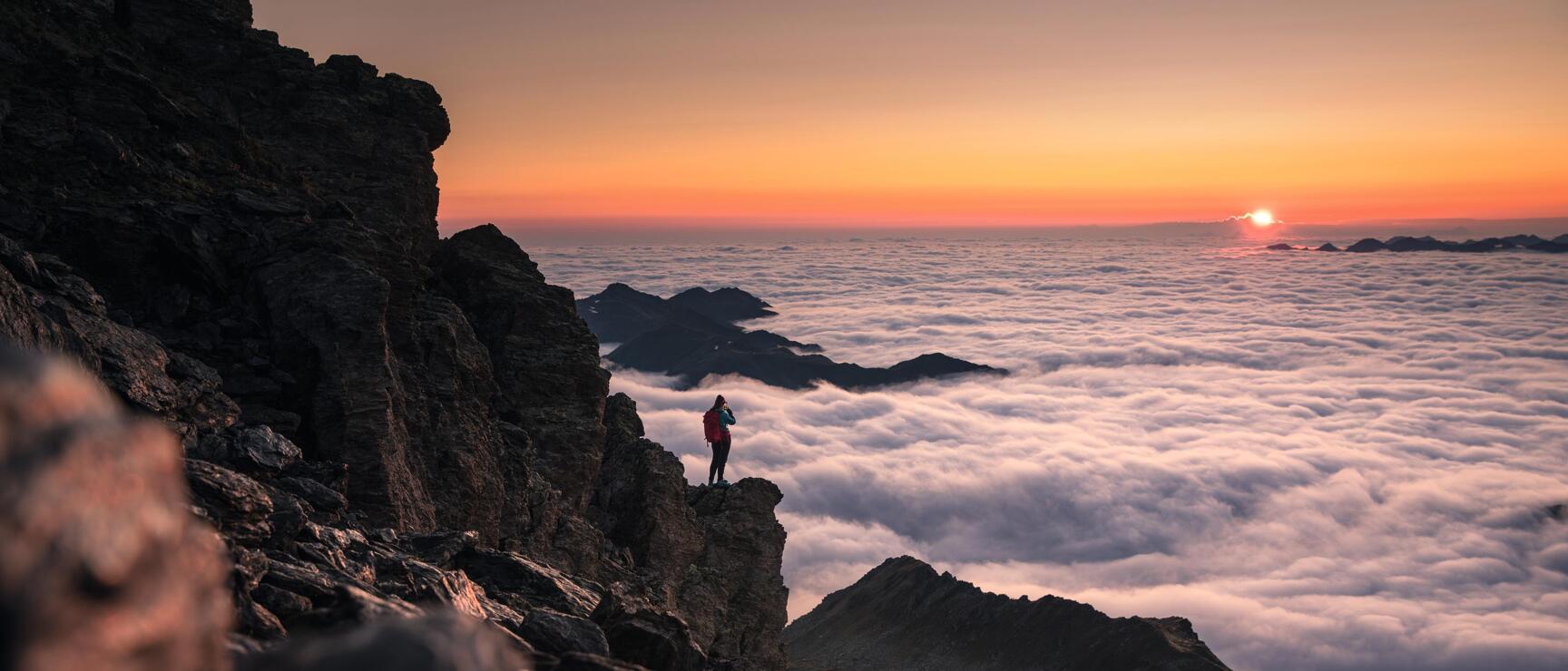 Person with red backpack on rock ledge above sea of clouds at orange sunset.
