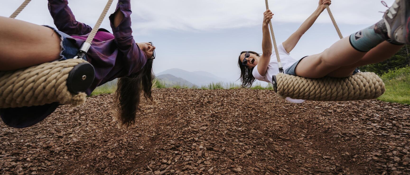 Two laughing women on rope swings with mountain panorama in background, worm's eye view.