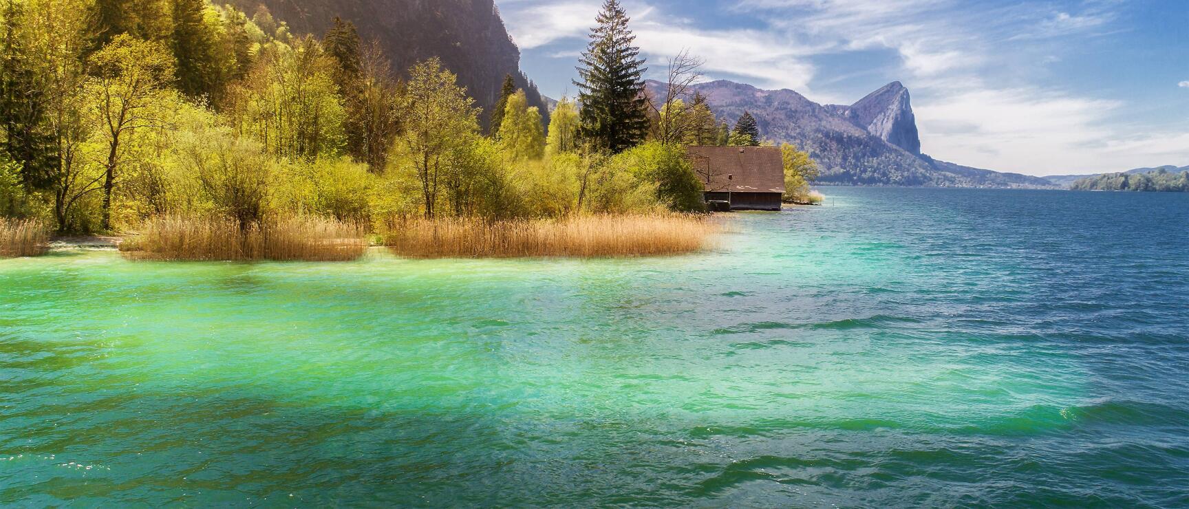 Holzhütte auf einer Halbinsel im türkisblauen Mondsee, Schilf, Bäume und Berge im Hintergrund.