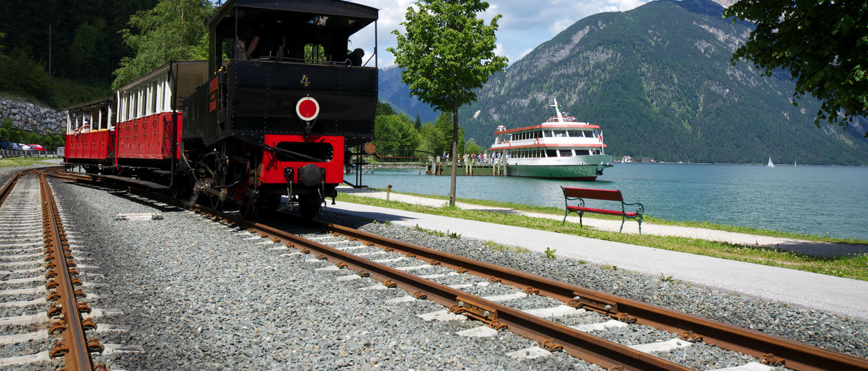 Historic steam locomotive with red carriages at Lake Achensee, excursion boat at dock, mountains in background.