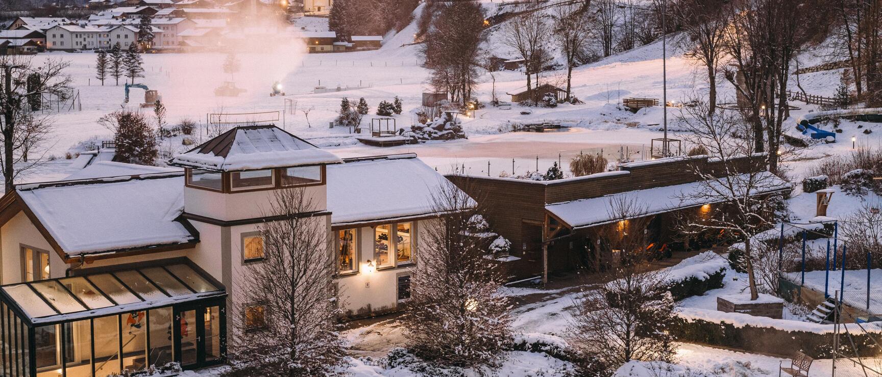 Heated outdoor pool of a hotel in winter, snow-covered Alpine village with church in the background, dusk.