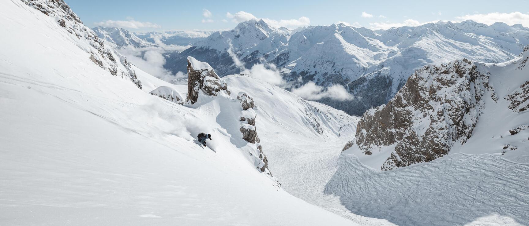 Skier in deep snow between rocks with panoramic snowy mountain peaks in the background.