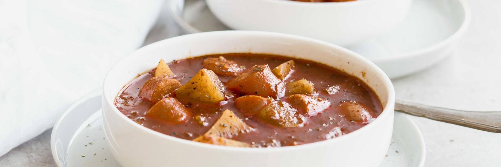 Two white bowls of goulash soup with potato and paprika pieces, spoon beside.