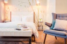Bright hotel room with white double bed, grey armchair, and wall mural with branches and birds.