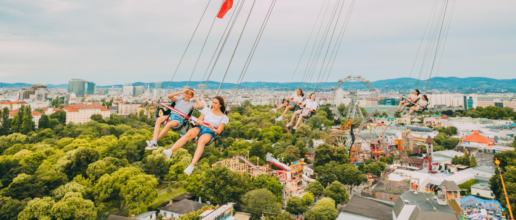 Personen im Kettenkarussell des Wiener Praters hoch über der Stadt, Riesenrad und Stadtpanorama im Hintergrund.