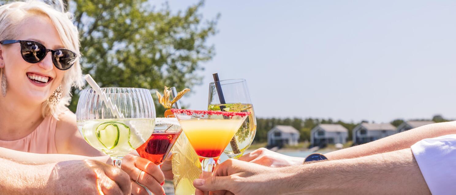 Multiple hands toasting with colourful cocktails, laughing woman with sunglasses, lake in the background.