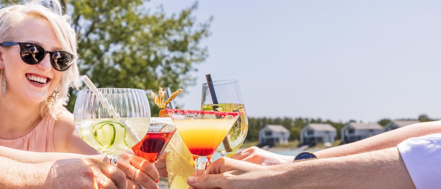 Multiple hands toasting with colourful cocktails, laughing woman with sunglasses, lake in the background.