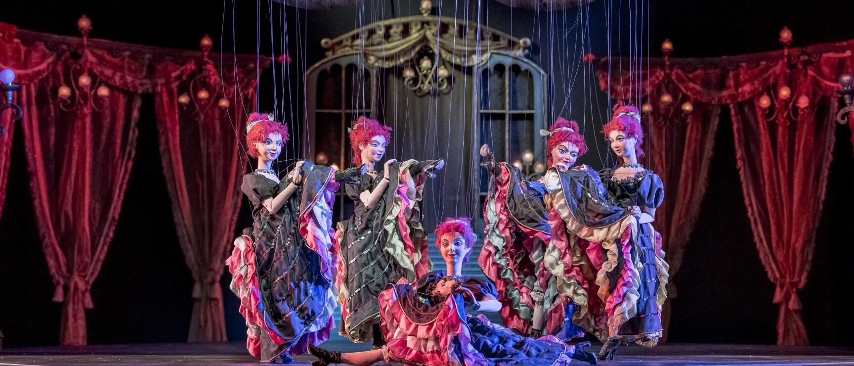 Five puppets in colorful ruffled dresses on illuminated stage with red curtains and bat decorations