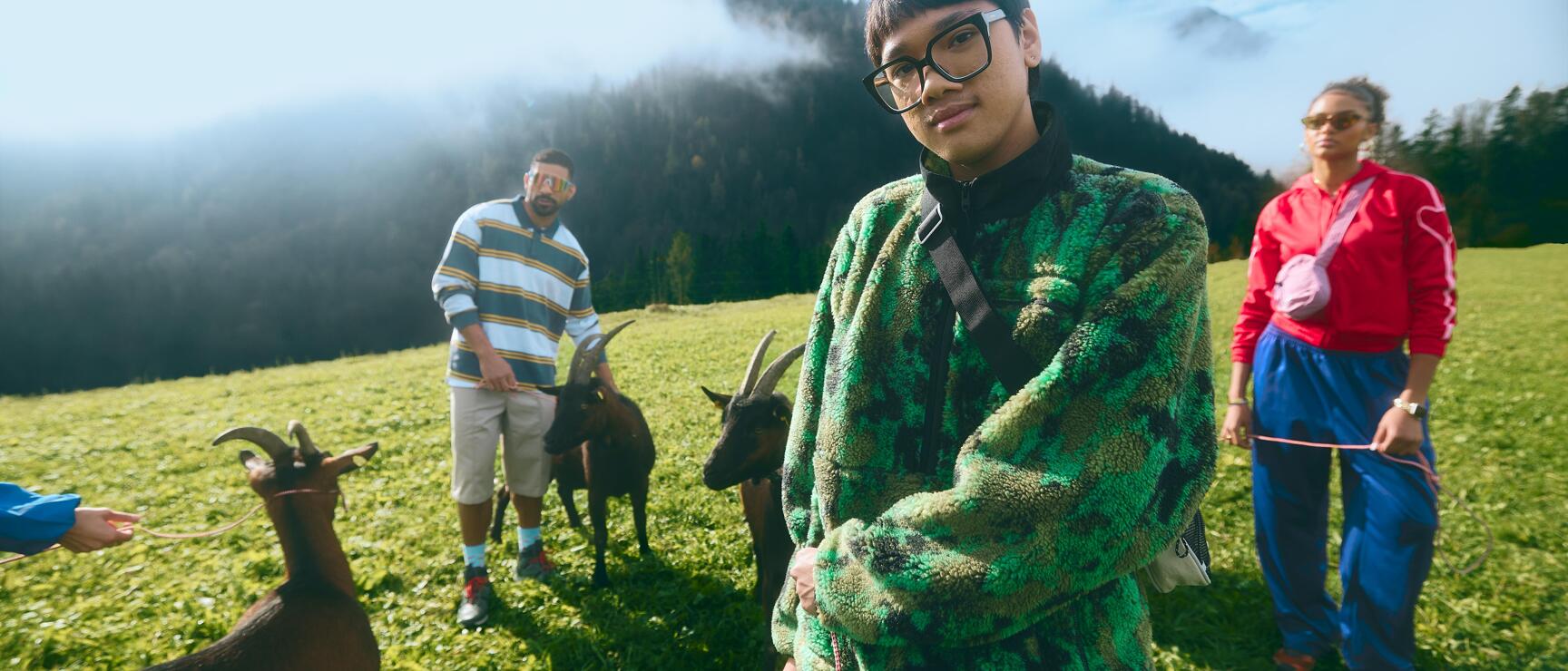 Three people with goats on an alpine meadow, forested mountains with fog in the background.