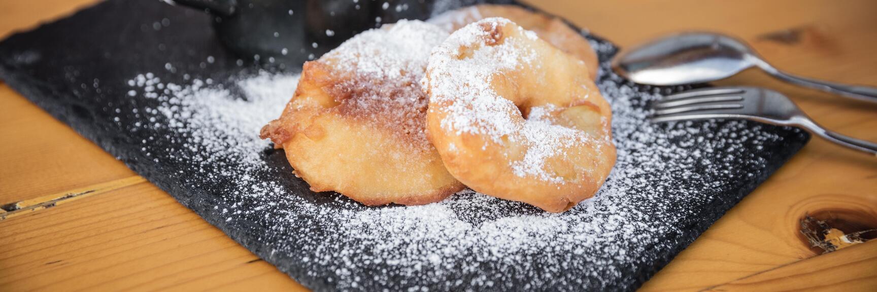 Two apple fritters dusted with icing sugar on a slate plate, small black pot, cutlery, wooden table.