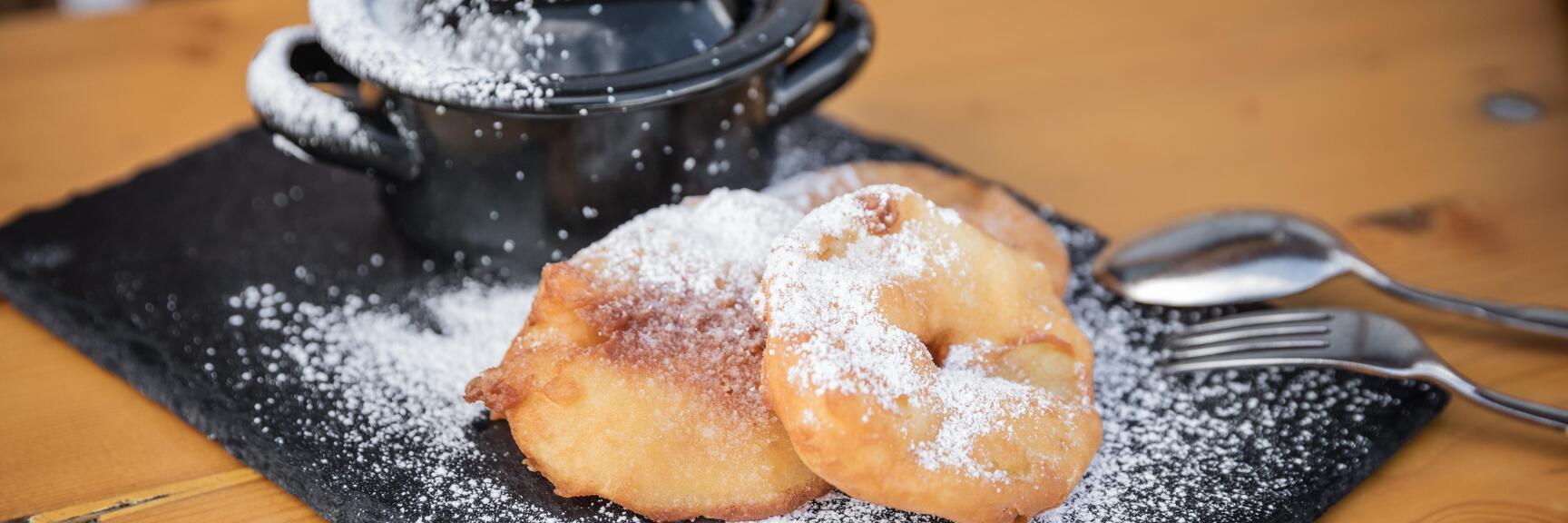 Two apple fritters dusted with icing sugar on a slate plate, small black pot, cutlery, wooden table.