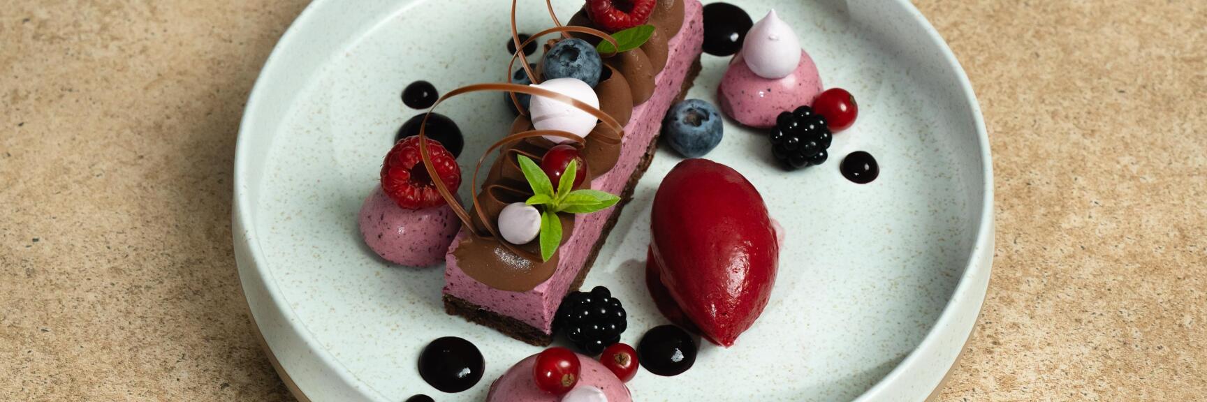 Dessert plate with berry mousse slice, red sorbet, fresh berries and chocolate decoration on light ceramic plate.