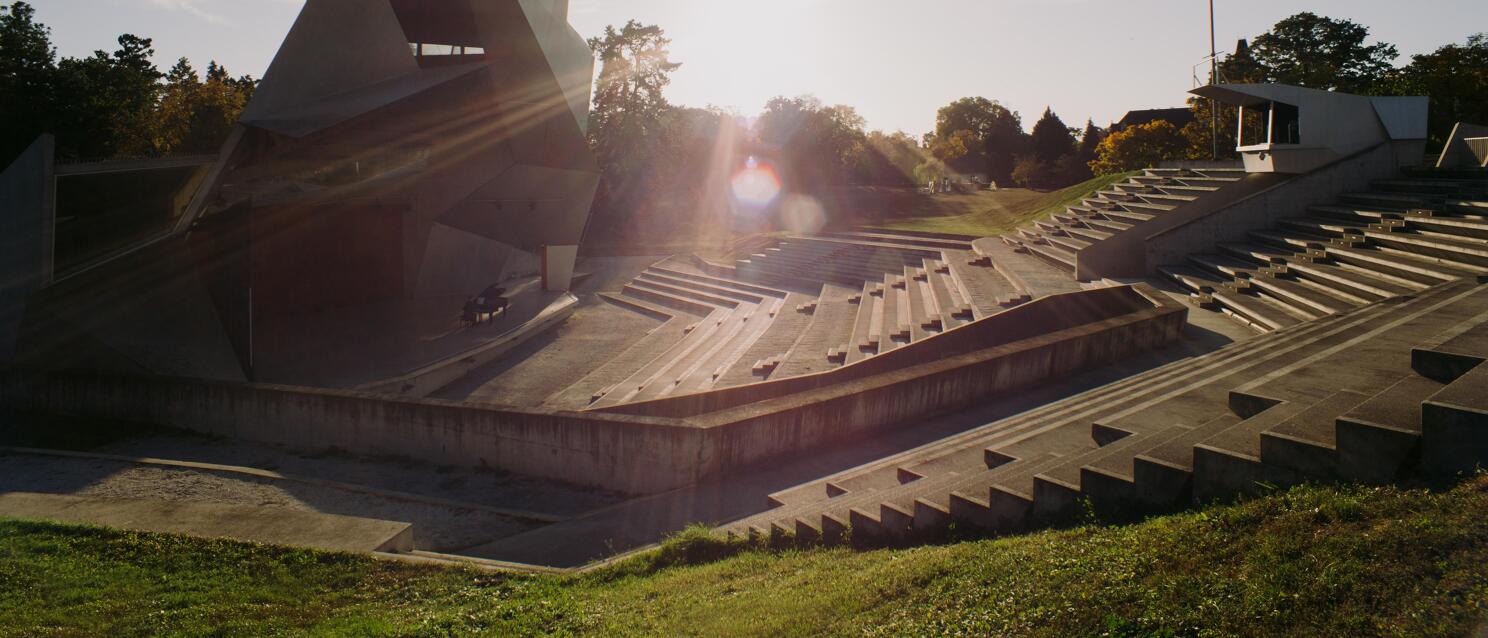 Concrete open-air theatre with distinctive geometric stage roof, stepped seating, trees backlit.