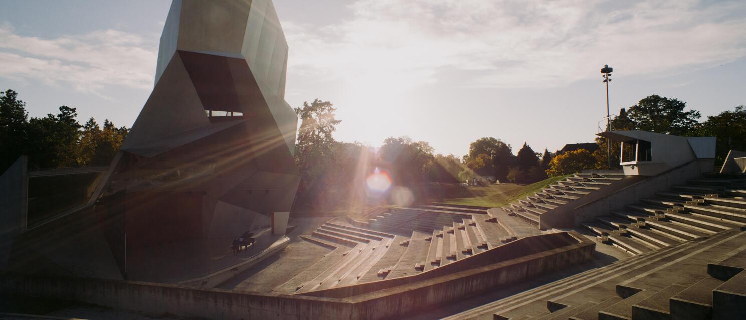 Concrete open-air theatre with distinctive geometric stage roof, stepped seating, trees backlit.
