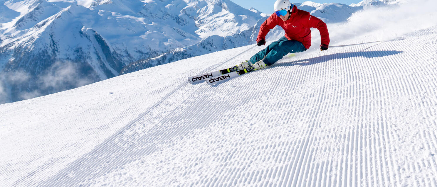 Skier in red jacket carving on prepared slope, snow-covered mountain peaks in background.