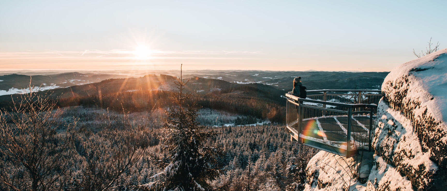 Person on snow-covered viewpoint above winter forest, sunset over hilly landscape.