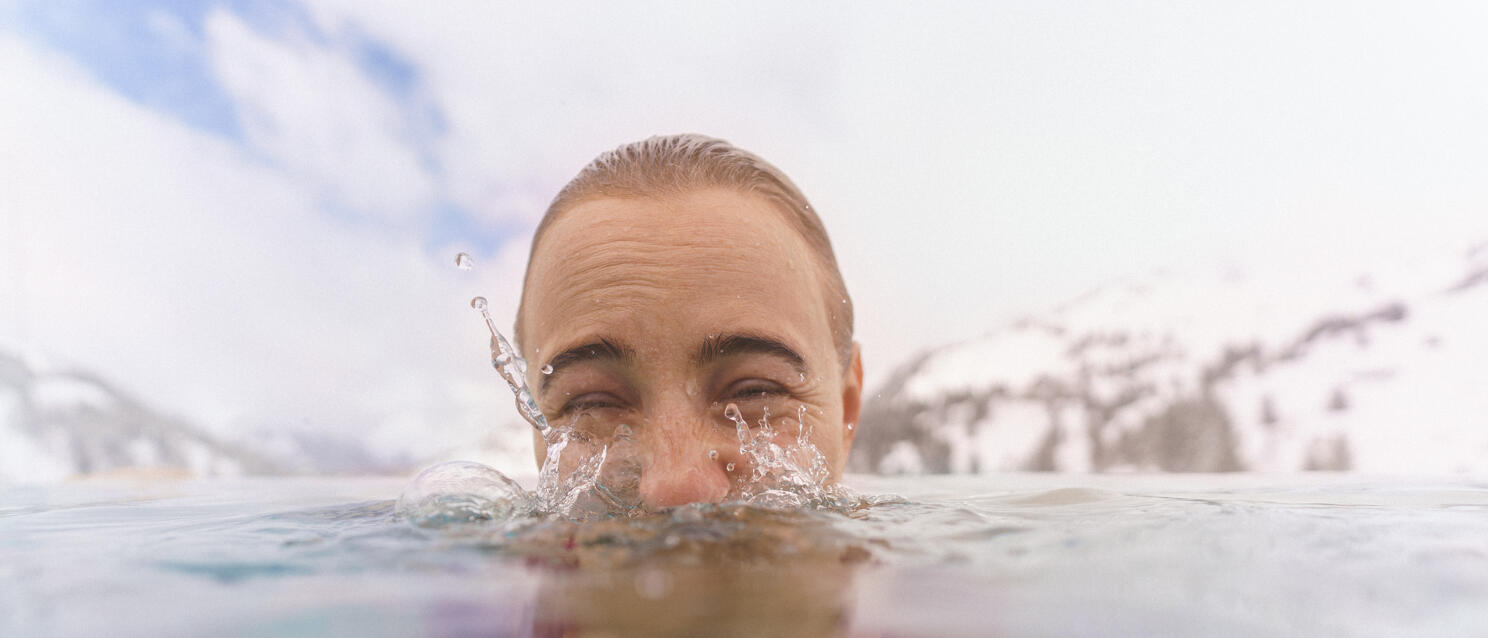 Woman in red bikini emerging from icy mountain lake with snow-covered peaks in background.