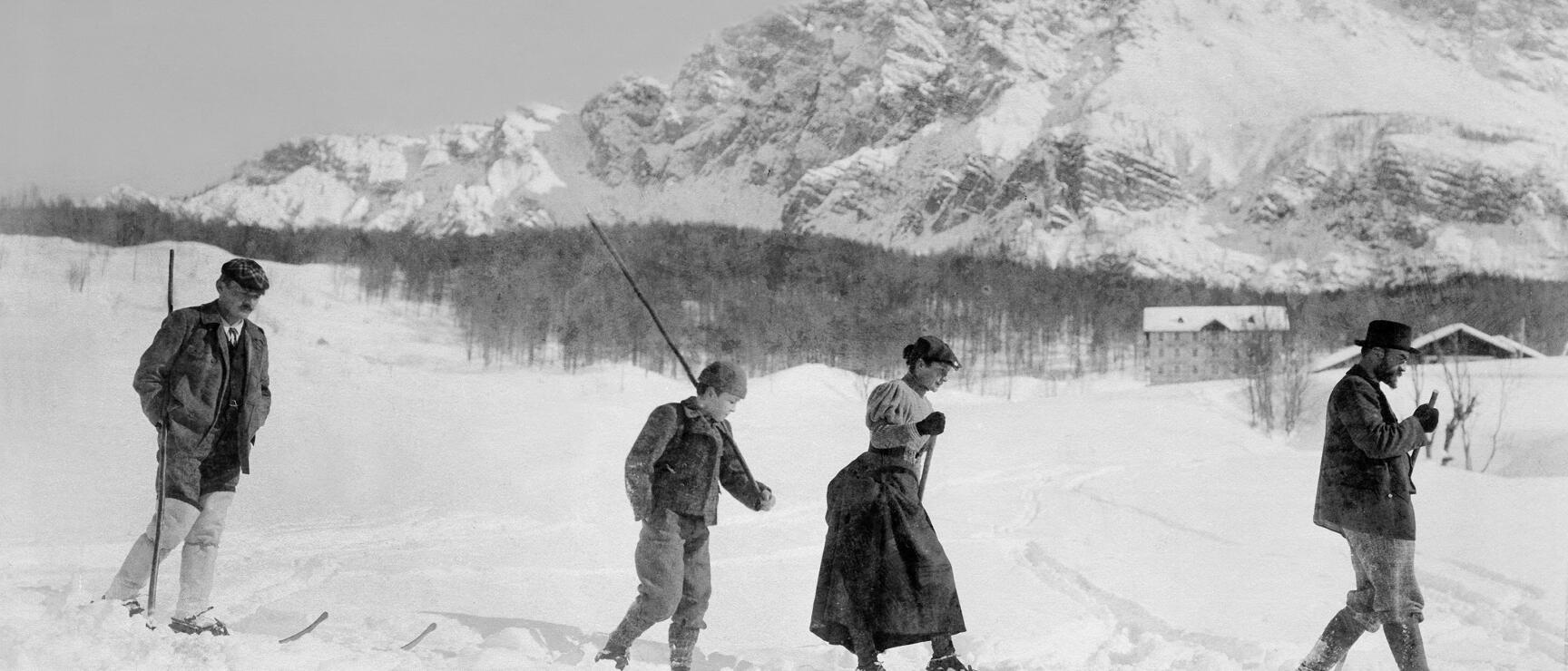 Historic black and white photo: Four people on skis in winter landscape, snow-covered mountains in background.