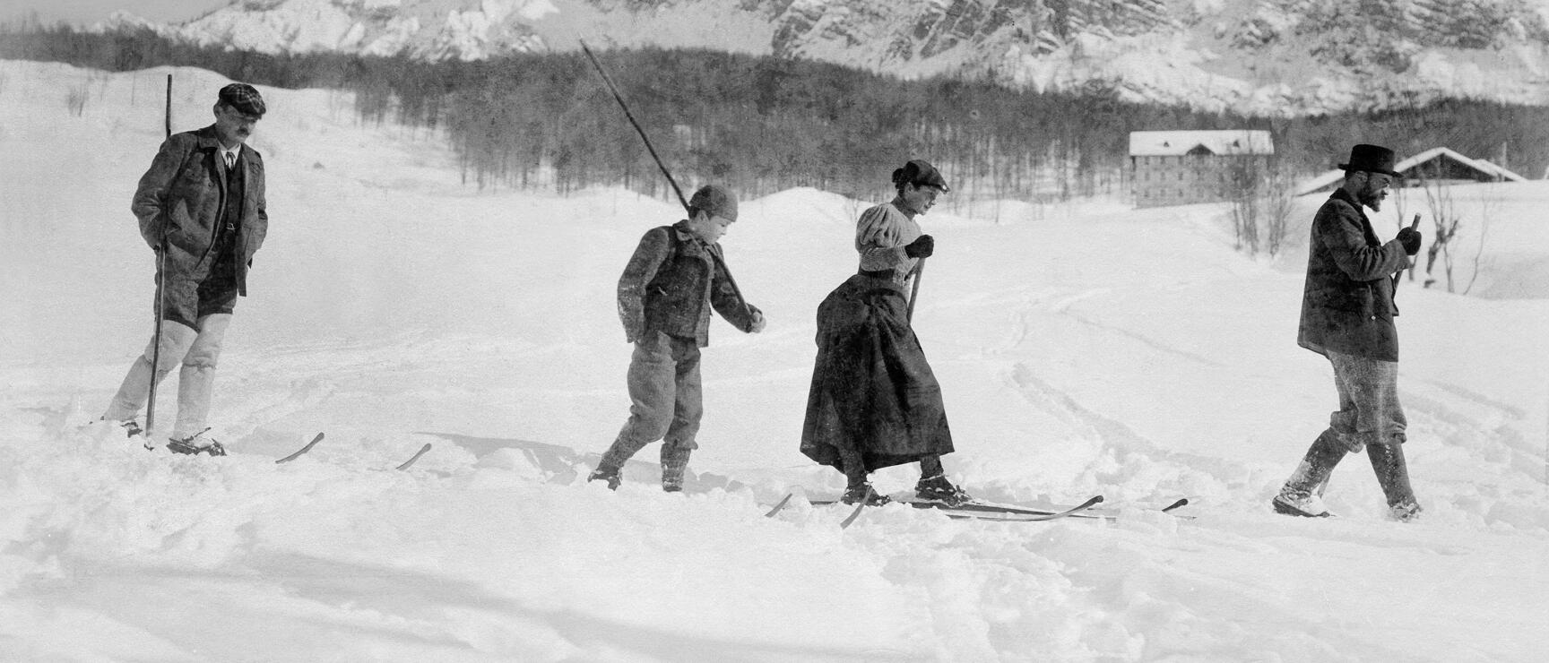 Historic black and white photo: Four people on skis in winter landscape, snow-covered mountains in background.
