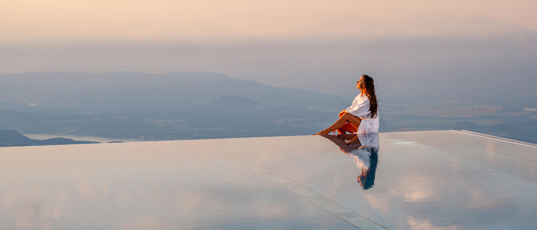 Woman in white bathrobe sitting at the edge of an infinity pool with panoramic views of hills and lake.
