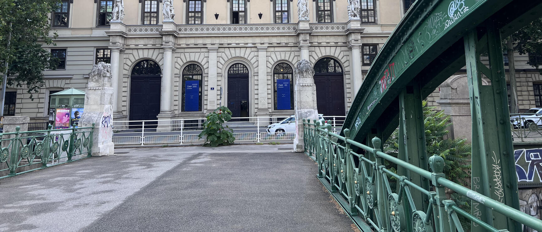 Historic building with sculptures and arches, green iron bridge with graffiti in foreground, Vienna.