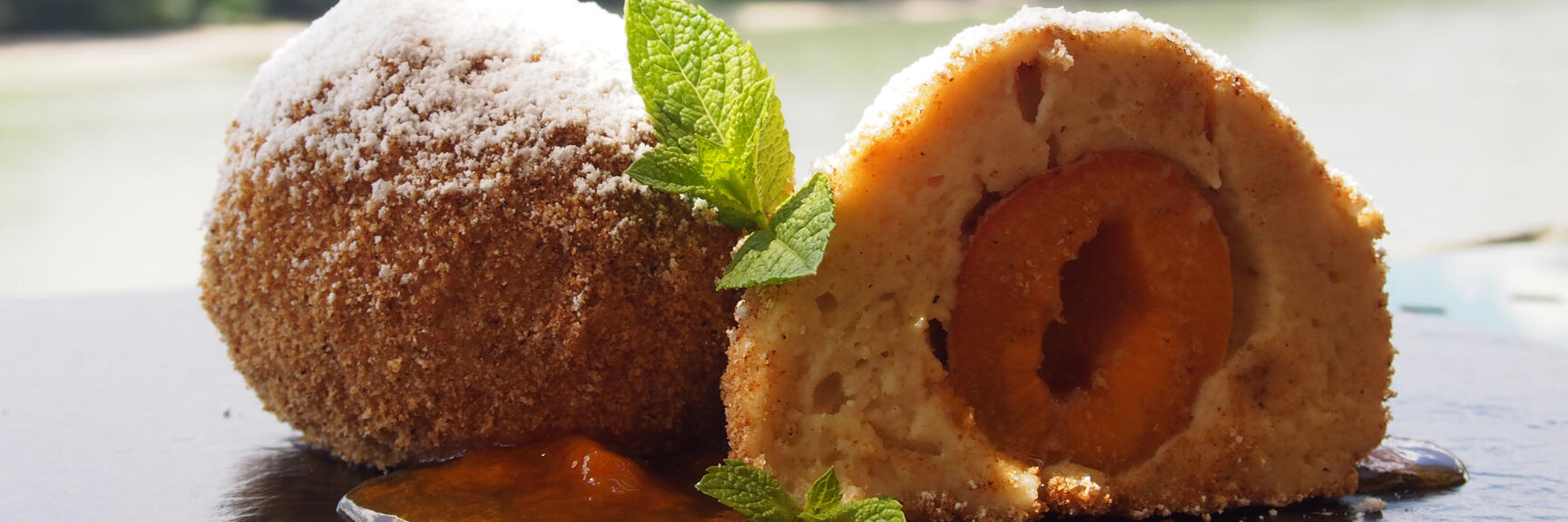 Two apricot dumplings with breadcrumbs and powdered sugar, one cut open, mint leaf, lake in background.