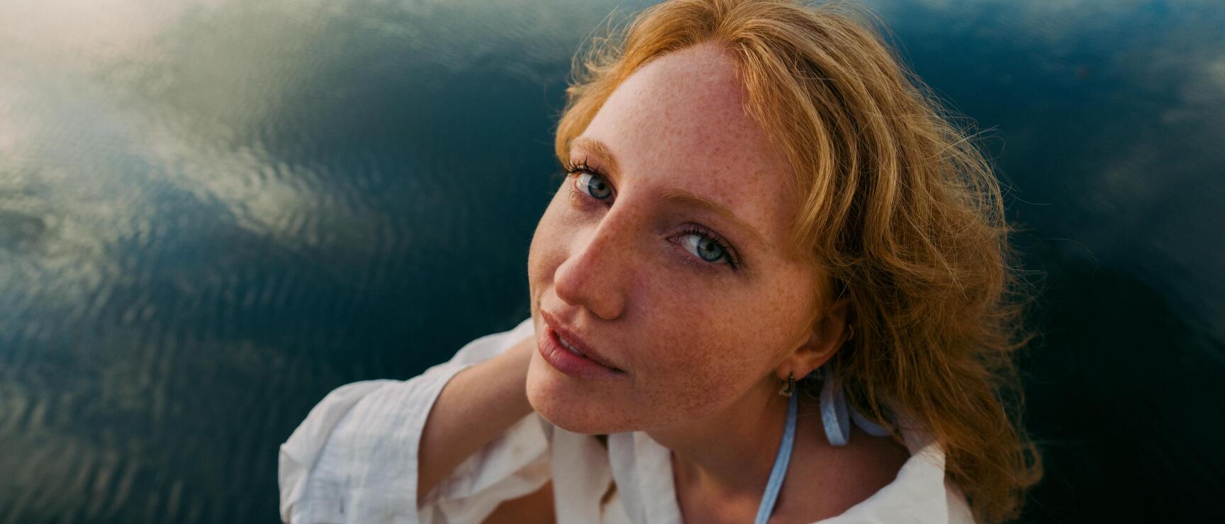 Young woman with red curls and white blouse sitting on a wooden jetty, dark lake water in the background.