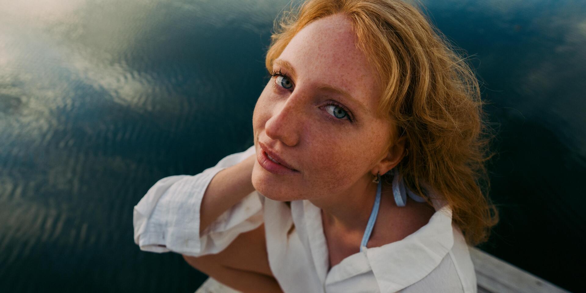 Young woman with red curls and white blouse sitting on a wooden jetty, dark lake water in the background.