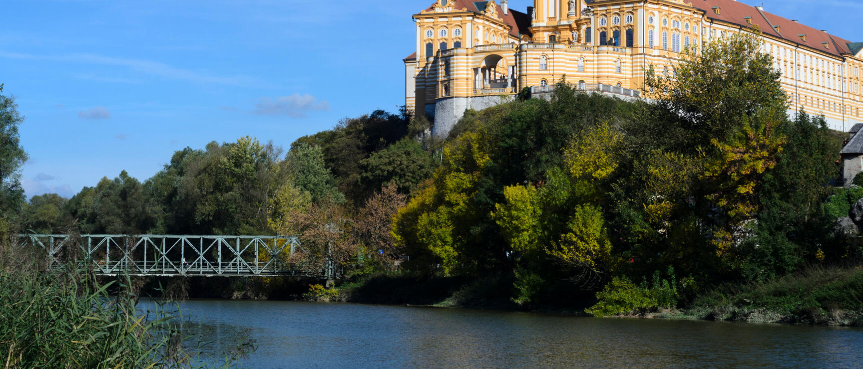 Melk Abbey on a rocky outcrop above the Danube, yellow Baroque facade with two towers, metal bridge in foreground.