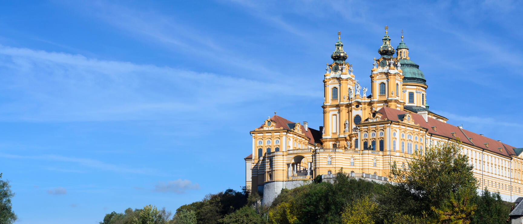 Stift Melk auf einem Felsen über der Donau, gelbe Barockfassade mit zwei Türmen, Metallbrücke im Vordergrund.