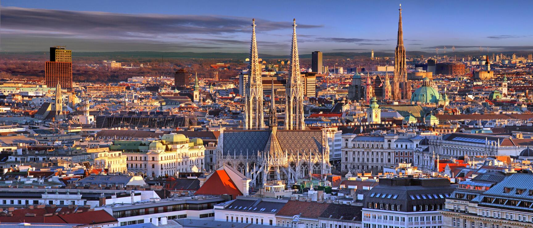 Panoramic view of Vienna with St Stephen's Cathedral and Votiv Church, old town roofs and blue evening sky.