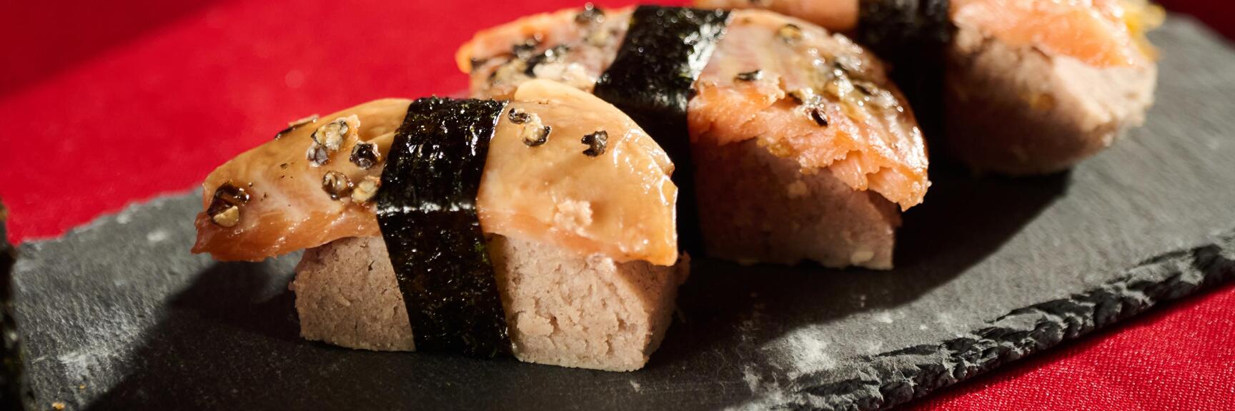 Vier Sushi-Stücke mit Nori und Sesam auf Schieferplatte, Sojasoße und Teetasse auf rotem Tisch.