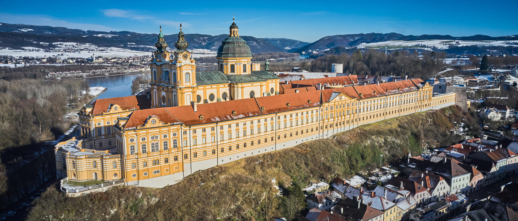 Aerial view of Melk Abbey on rocky outcrop above Danube, yellow Baroque façade, red roof, winter landscape.