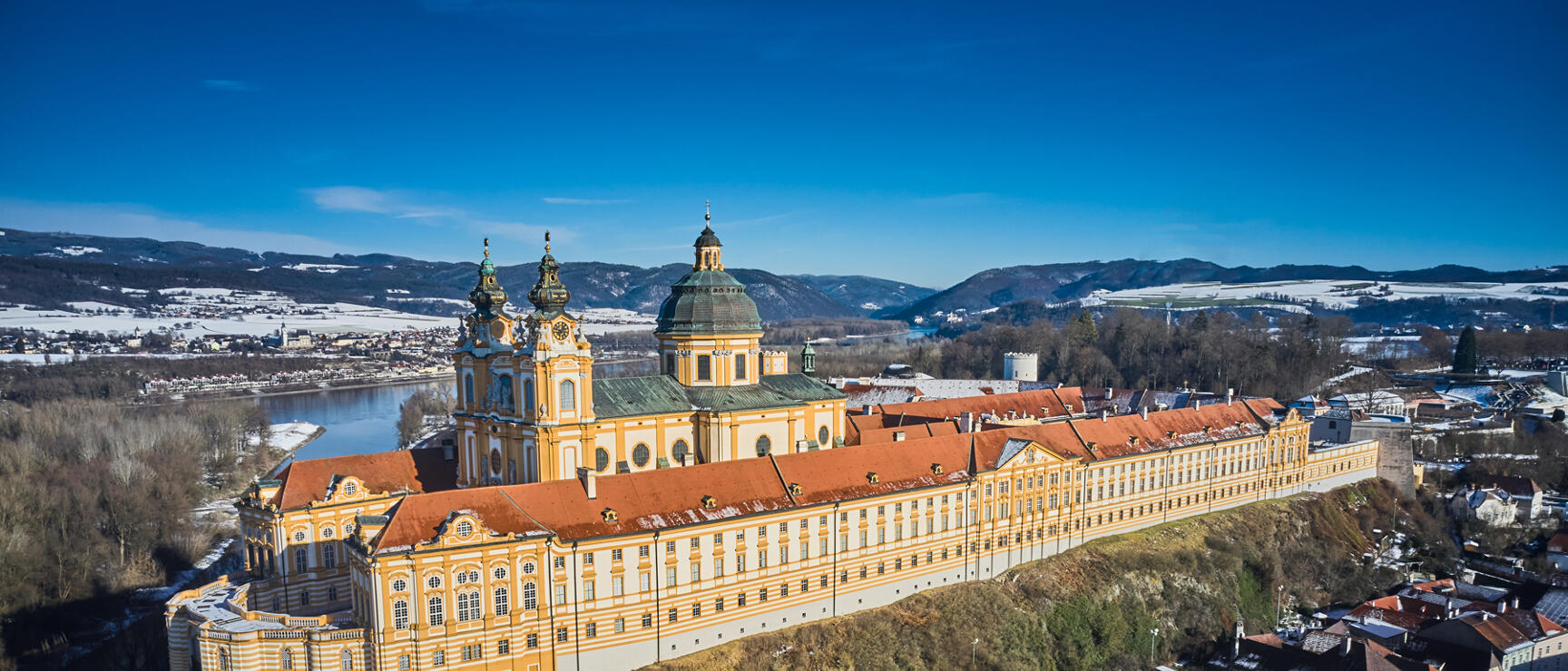 Aerial view of Melk Abbey on rocky outcrop above Danube, yellow Baroque façade, red roof, winter landscape.