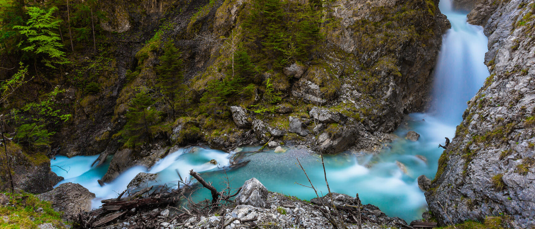 Tall waterfall cascading into a rocky gorge with turquoise water, moss-covered rock walls and coniferous trees.