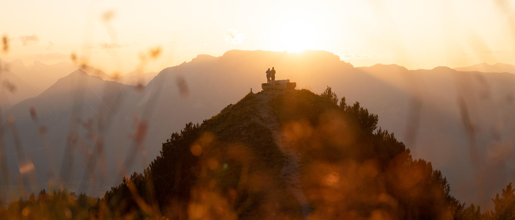 Two people silhouetted on a mountain peak at sunset with Alpine panorama in the background.