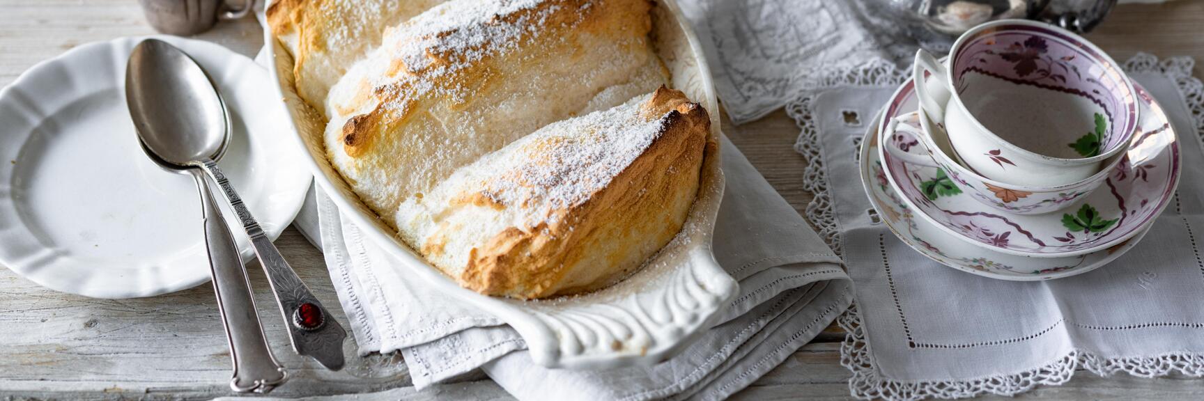 Salzburger Nockerl mit Puderzucker in weißer Auflaufform, daneben Porzellangeschirr und Silberkanne.
