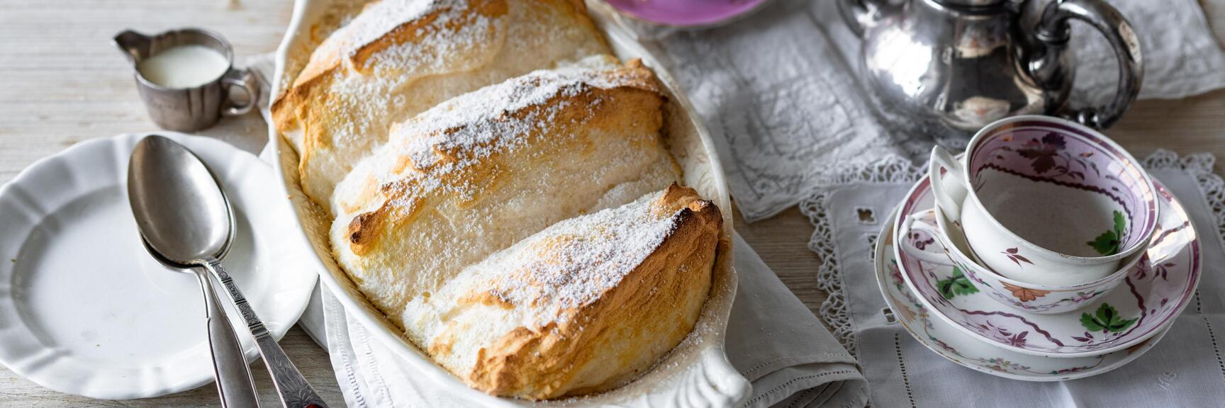 Salzburg dumplings dusted with powdered sugar in a white baking dish, with porcelain tableware and a silver jug beside it.