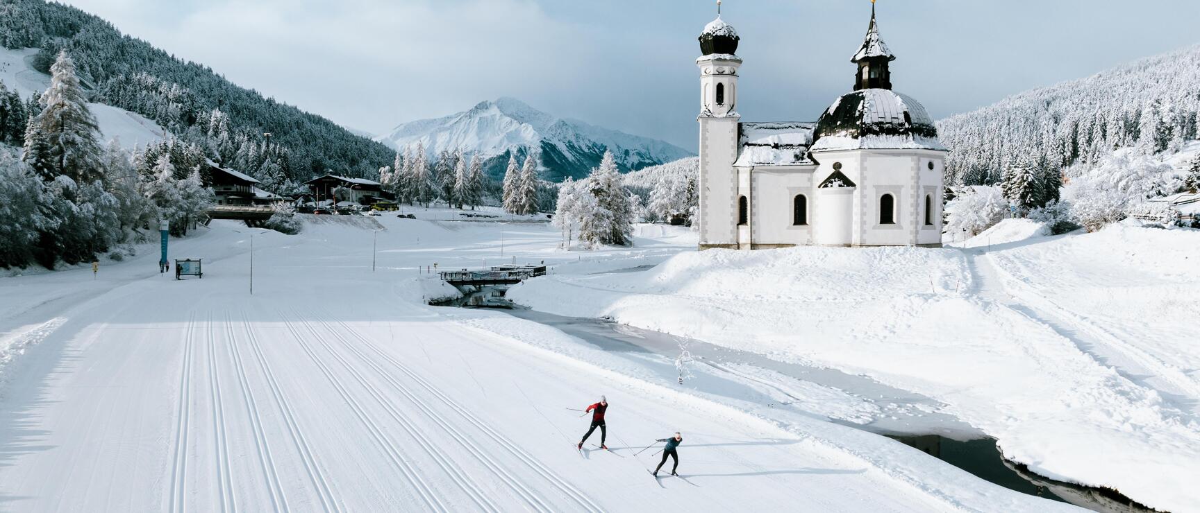 Two cross-country skiers on prepared track next to white Baroque church, snowy mountains in background, Seefeld.