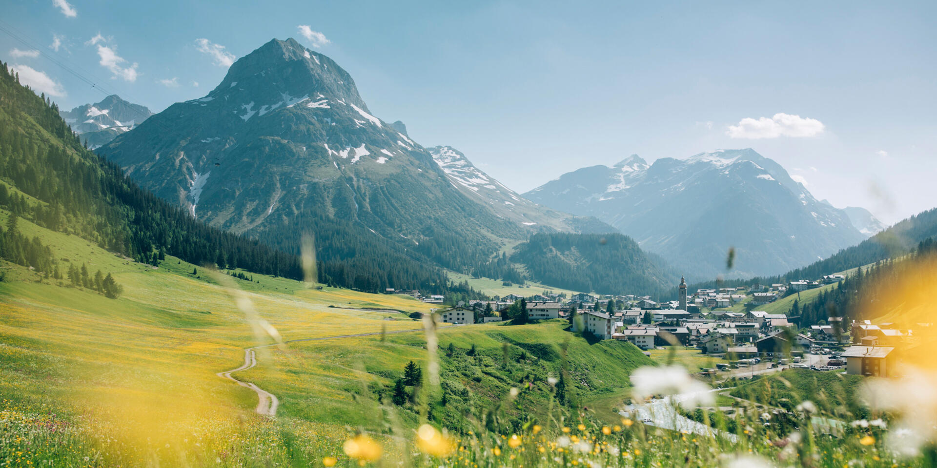 Blooming alpine meadow with path, view of Lech at Arlberg, snow-capped mountain peaks in background.