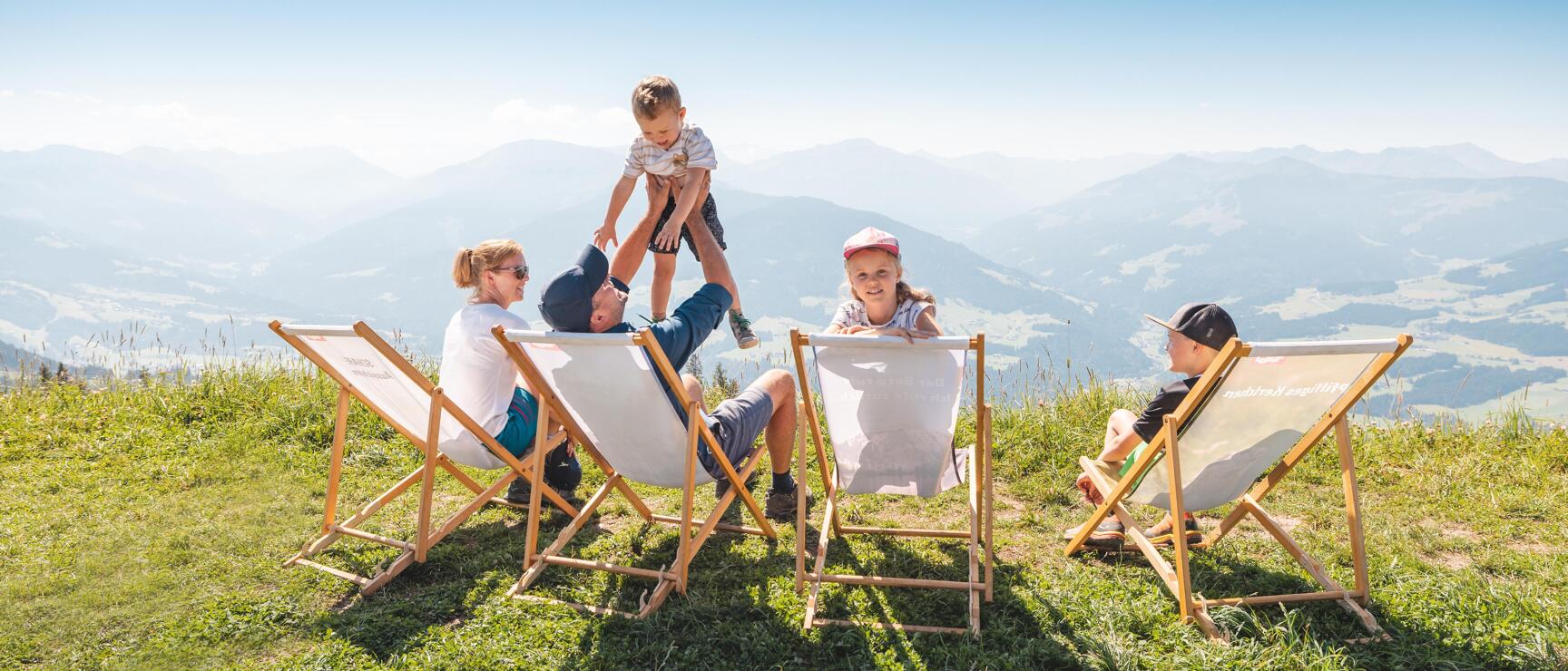 Two adults and two children in deckchairs on an alpine meadow with mountain panorama in the background.