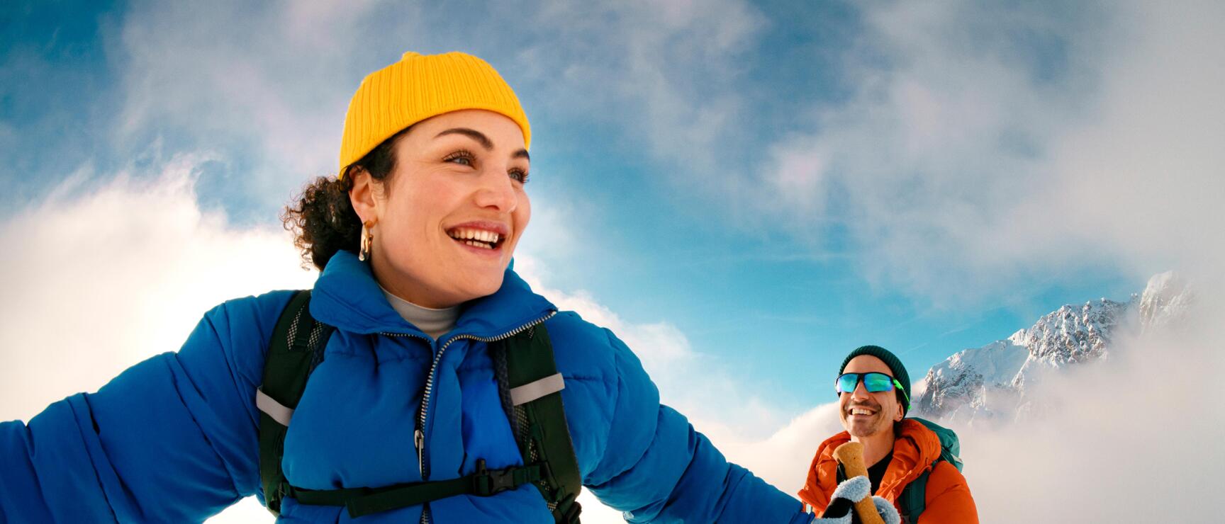 Two snowshoers are laughing as they make their way through the snow-covered mountain landscape of the Austrian Alps. The vibrant colors of their winter clothing, the soft clouds, and the vast snowy backdrop convey a sense of joy, lightheartedness, and the shared experience of an active winter day in nature.