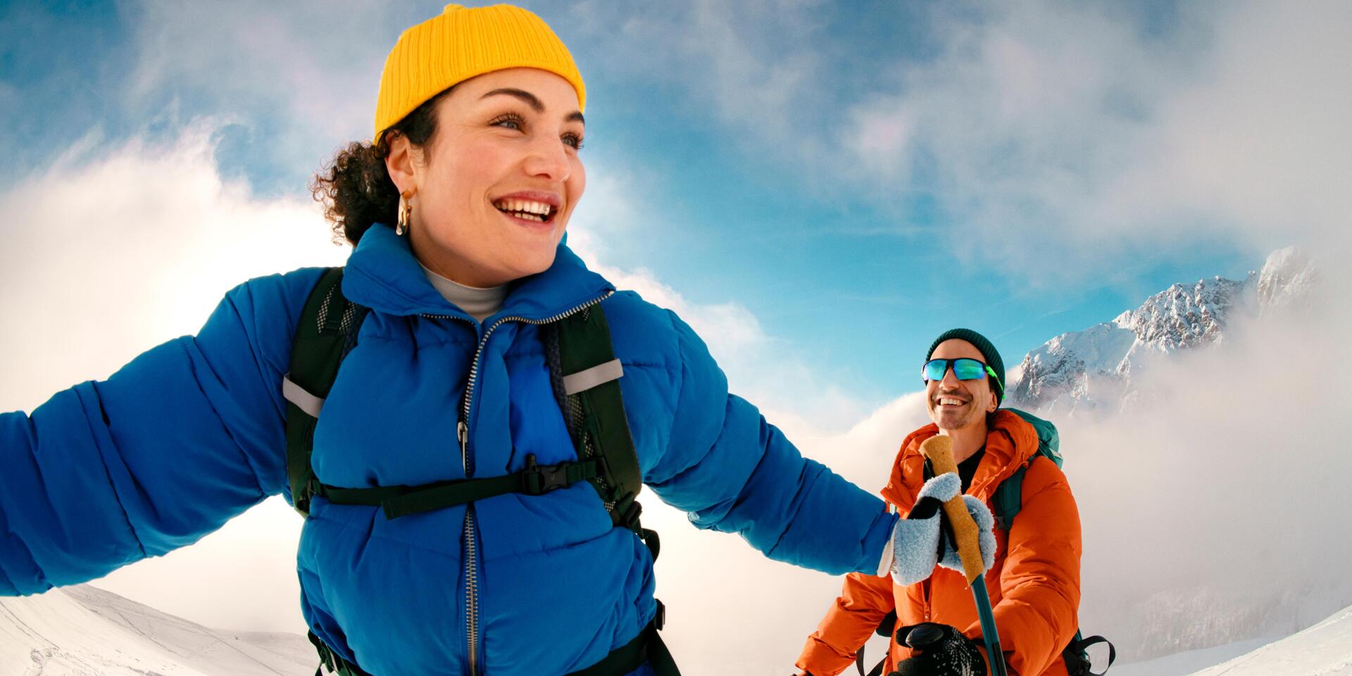 Two snowshoers are laughing as they make their way through the snow-covered mountain landscape of the Austrian Alps. The vibrant colors of their winter clothing, the soft clouds, and the vast snowy backdrop convey a sense of joy, lightheartedness, and the shared experience of an active winter day in nature.